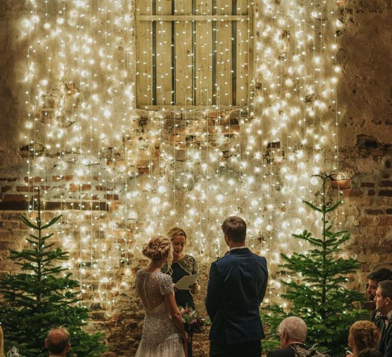 Bride and groom tie the knot at winter ceremony wearing a embellished wedding dress and navy velvet suit with fairy light curtain and Christmas trees