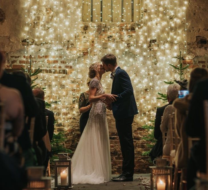 Bride and groom tie the knot at winter ceremony wearing a embellished wedding dress and navy velvet suit with fairy light curtain and Christmas trees