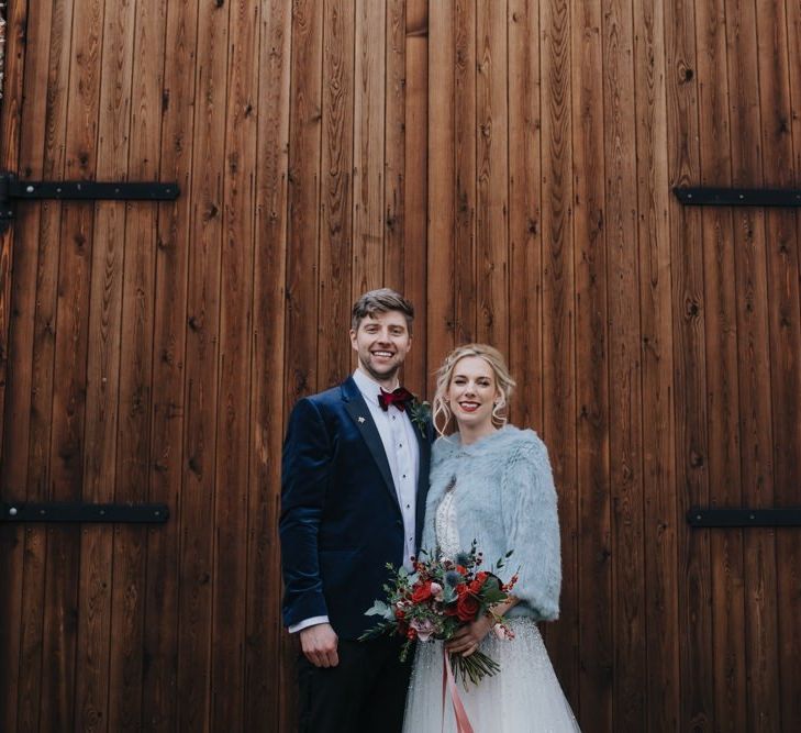 Bride wearing embellished wedding dress and jacket at winter wedding ceremony with red floral bouquet