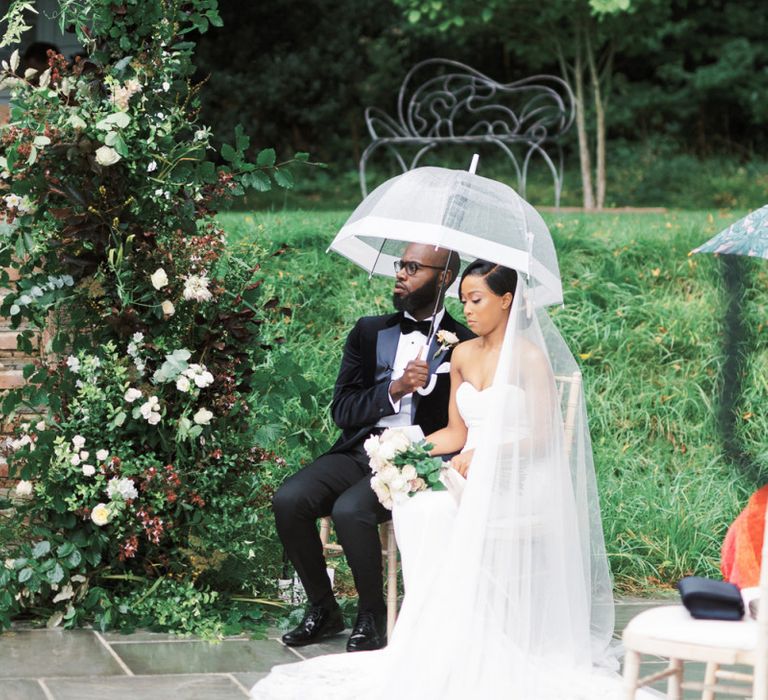 Bride and groom sitting under a clear umbrella during the outdoor wedding ceremony