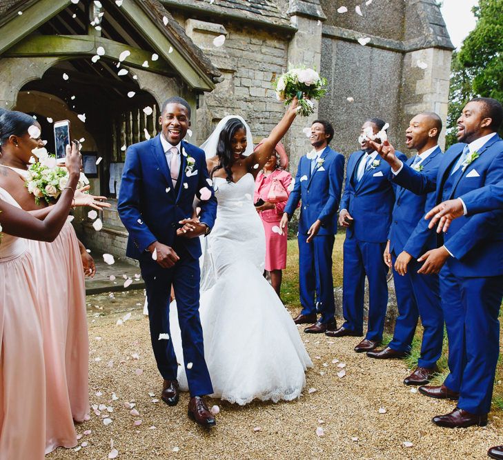 Bride in Lace Fishtail Gown with Strapless Sweetheart Neckline by Suzanne Neville | Lace Edged Cathedral Length Veil | Groom in Blue Three-Piece Ted Baker Suit with Blush Pink Tie | Bridal Bouquet of White and Blush Roses and Peonies | Bridesmaids in Blush Pink One Shoulder Dresses by Vera Wang | Groomsmen in  Blue Three-Piece Suits by Ted Baker | Confetti Throw at St John the Baptist Church, Elmore | Peg Board Table Plan &amp; DIY Sweet Table for an Elmore Court Wedding | Mavric Photography