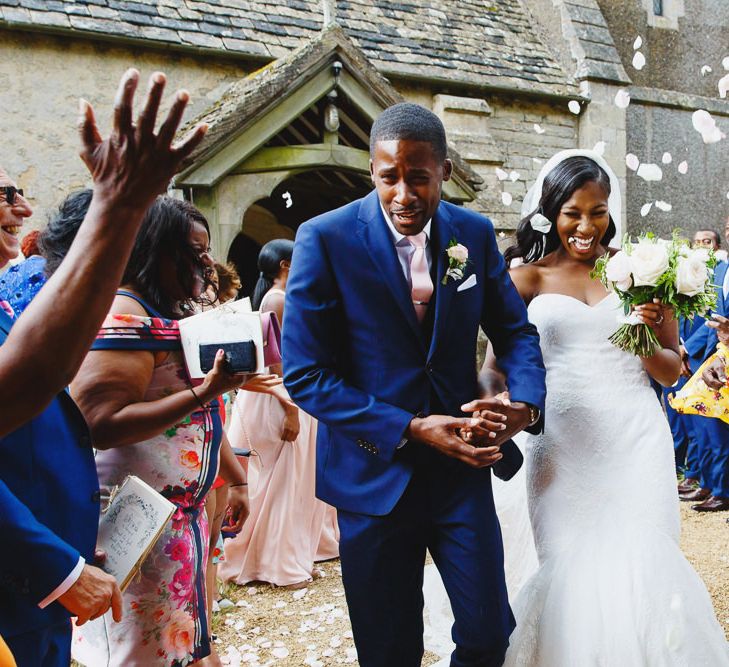 Bride in Lace Fishtail Gown with Strapless Sweetheart Neckline by Suzanne Neville | Lace Edged Cathedral Length Veil | Groom in Blue Three-Piece Ted Baker Suit with Blush Pink Tie | Bridal Bouquet of White and Blush Roses and Peonies | Confetti Throw at St John the Baptist Church, Elmore | Peg Board Table Plan &amp; DIY Sweet Table for an Elmore Court Wedding | Mavric Photography