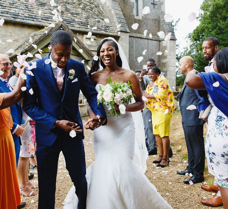 Bride in Lace Fishtail Gown with Strapless Sweetheart Neckline by Suzanne Neville | Lace Edged Cathedral Length Veil | Groom in Blue Three-Piece Ted Baker Suit with Blush Pink Tie | Bridal Bouquet of White and Blush Roses and Peonies | Confetti Throw at St John the Baptist Church, Elmore | Peg Board Table Plan &amp; DIY Sweet Table for an Elmore Court Wedding | Mavric Photography