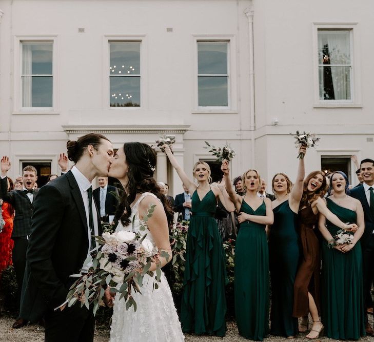 Bride and groom kiss in front of  guests and  bridesmaids wearing emerald green bridesmaid dresses