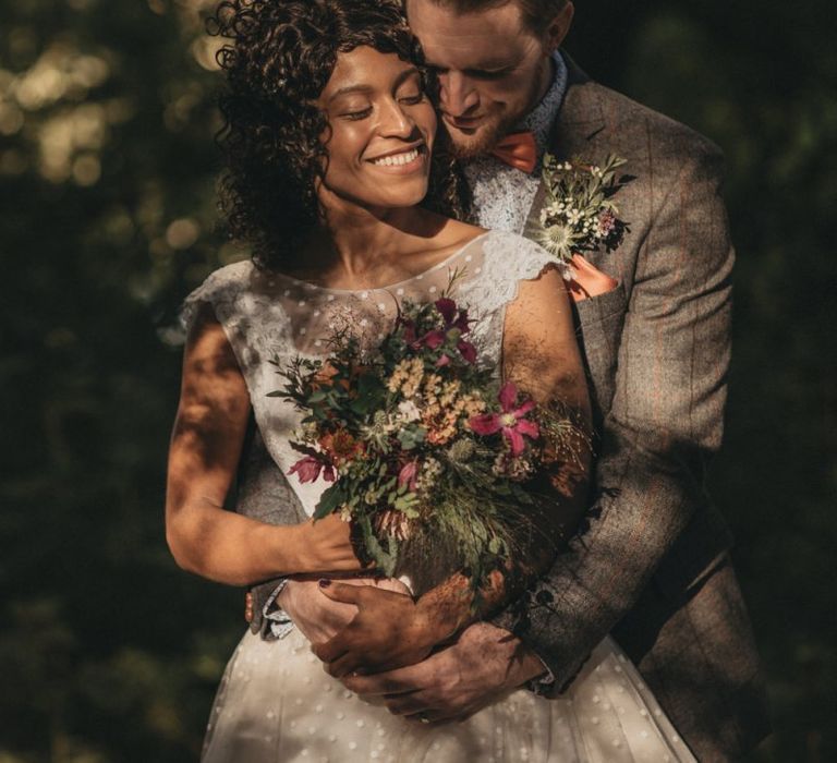 Groom in Grey Check Suit Embracing His Bride Holding a Wildflower Wedding Bouquet