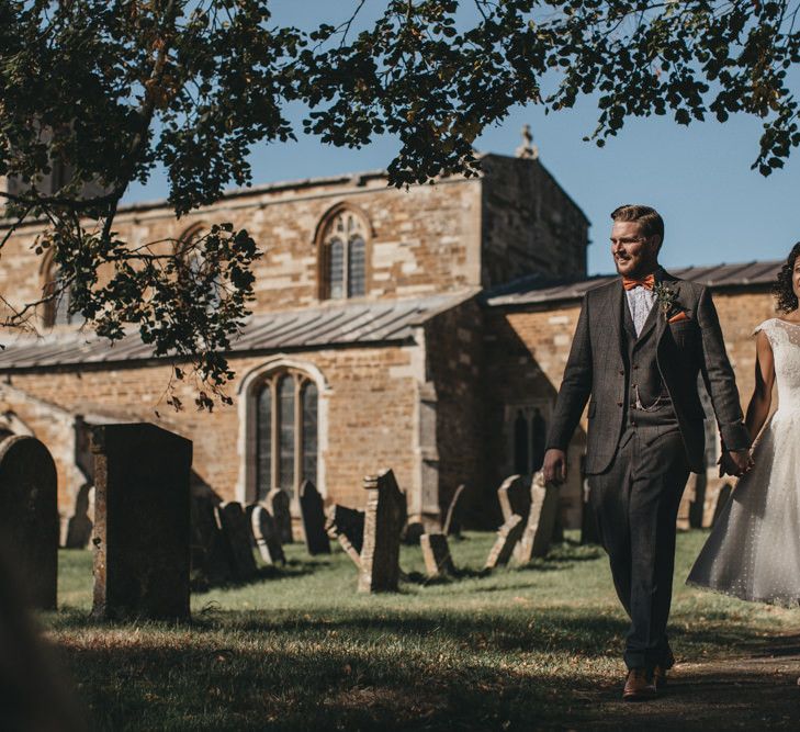 Bride in Polka Dot Tulle Wedding Dress and Groom in Grey Wool Suit Walking Through the Church Courtyard