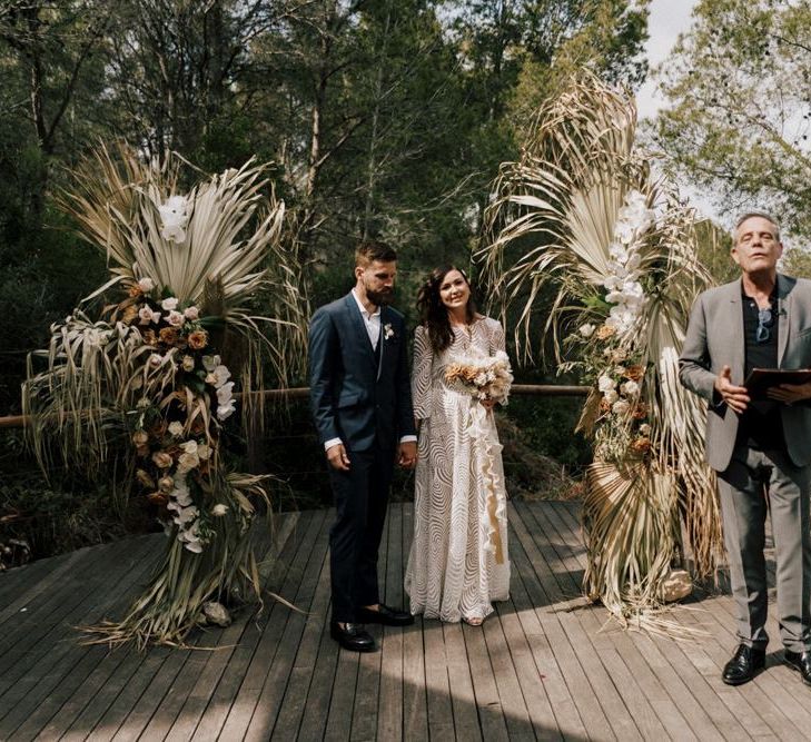 Bride and Groom Standing at the Floral Altar in a Margaux Tardits Wedding Dress and Moss Bros. Navy Wedding Suit