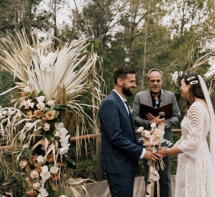 Bride in Margaux Tardits Wedding Dress  and Groom in a Navy Wedding Suit Holding Hands at the Altar