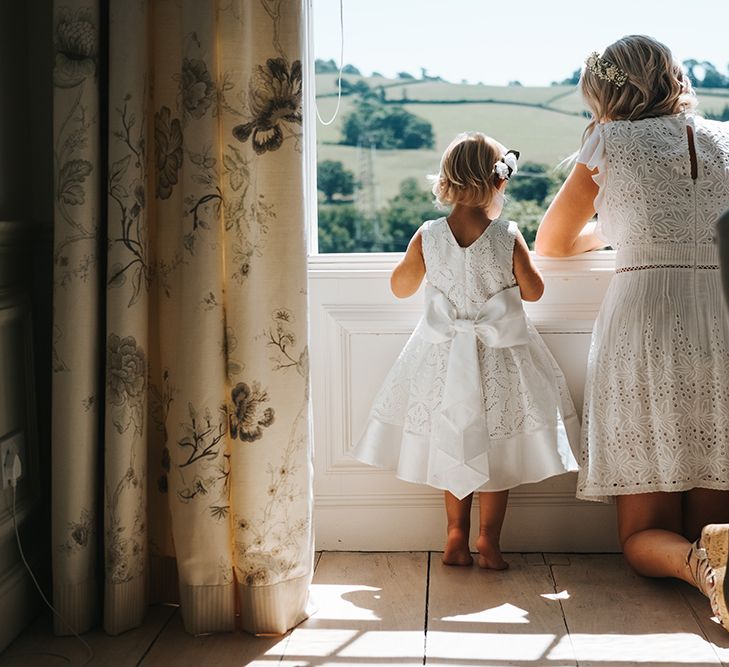 Bridesmaid in White Broderie Anglaise Dress Crouching Down Looking out The Window with a Little Flower Girl