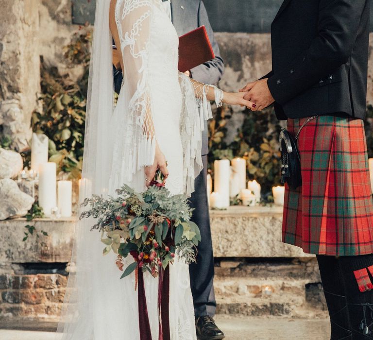 Bride in Bohemian Rue De Seine Wedding Dress and Groom in Red Tartan Kilt Holding Hands During The Wedding Ceremony