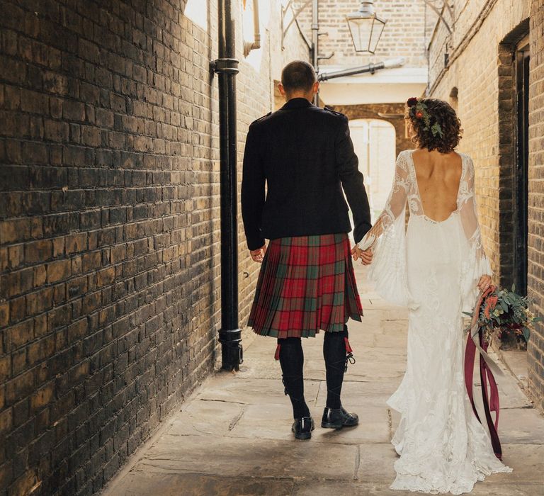 Bride in Lace Rue De Seine Wedding Dress  with Tassel Bell Sleeves Low Back and Groom in Red Tartan Kilt Highland Wear Holding Hands