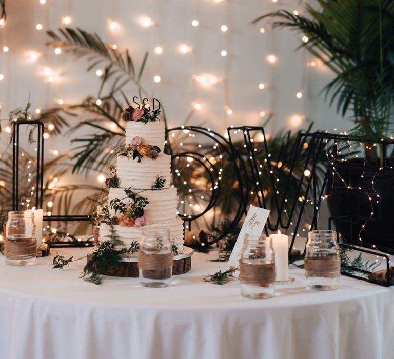 Four Tier Buttercream Wedding Cake with Foliage Decor Standing in Front of Fairy Light Curtain