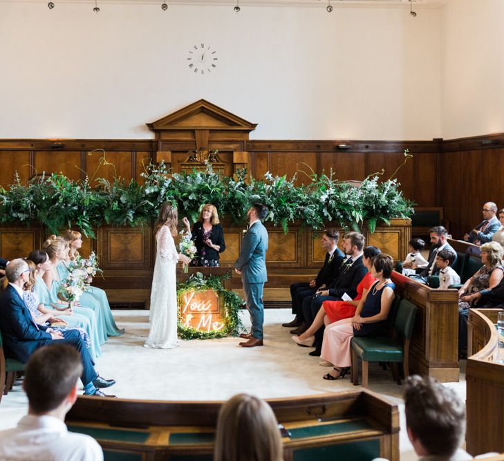 Bride in Grace Loves Lace Verdelle Dress with V-Neck, Low Square Back, Mid-Thigh Front Split and Lace Cape | Foliage Hair Crown | Pastel Bridal Bouquet | Groom in Blue Reiss Suit with Brown Grenson Shoes | Neon You &amp; Me Sign at Altar | Bridesmaids in Mismatched Marine Green Rewritten Dresses | Town Hall Hotel Bethnal Green | Neon Sign and Marine Green Rewritten Bridesmaids Dresses with Lace Cape Grace Loves Lace Gown | Alain Mbouche Photography