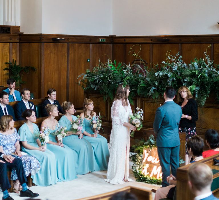 Bride in Grace Loves Lace Verdelle Dress with V-Neck, Low Square Back, Mid-Thigh Front Split and Lace Cape | Foliage Hair Crown | Pastel Bridal Bouquet | Groom in Blue Reiss Suit with Brown Grenson Shoes | Neon You &amp; Me Sign at Altar | Bridesmaids in Mismatched Marine Green Rewritten Dresses | Town Hall Hotel Bethnal Green | Neon Sign and Marine Green Rewritten Bridesmaids Dresses with Lace Cape Grace Loves Lace Gown | Alain Mbouche Photography