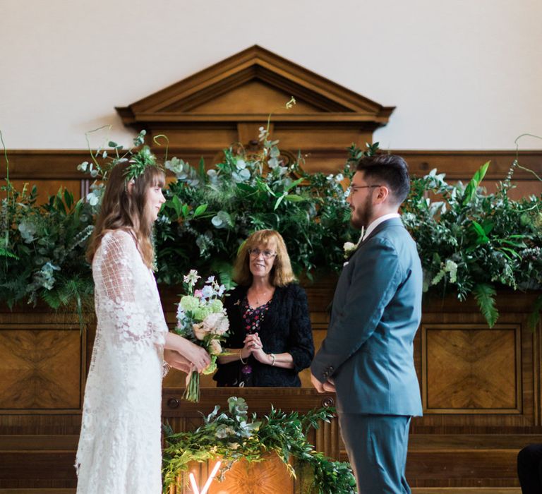 Bride in Grace Loves Lace Verdelle Dress with V-Neck, Low Square Back, Mid-Thigh Front Split and Lace Cape | Foliage Hair Crown | Pastel Bridal Bouquet | Groom in Blue Reiss Suit with Brown Grenson Shoes | Neon You &amp; Me Sign at Altar | Town Hall Hotel Bethnal Green | Neon Sign and Marine Green Rewritten Bridesmaids Dresses with Lace Cape Grace Loves Lace Gown | Alain Mbouche Photography