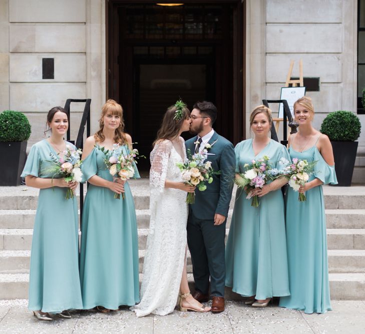 Bride in Grace Loves Lace Verdelle Dress with V-Neck, Low Square Back, Mid-Thigh Front Split and Lace Cape | Foliage Hair Crown | Gold Flat Gianvito Rossi Sandals | Groom in Blue Reiss Suit with Brown Grenson Shoes | Bridesmaids in Mismatched Marine Green Rewritten Dresses | Pastel Wedding Bouquets | Neon Sign and Marine Green Rewritten Bridesmaids Dresses with Lace Cape Grace Loves Lace Gown | Alain Mbouche Photography