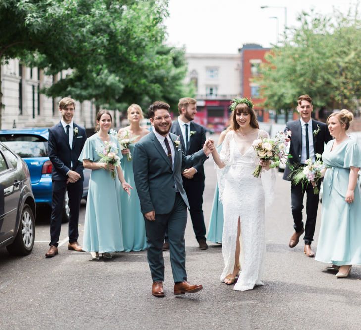 Bride in Grace Loves Lace Verdelle Dress with V-Neck, Low Square Back, Mid-Thigh Front Split and Lace Cape | Foliage Hair Crown | Gold Flat Gianvito Rossi Sandals | Groom in Blue Reiss Suit with Brown Grenson Shoes | Bridesmaids in Mismatched Marine Green Rewritten Dresses | Pastel Wedding Bouquets | Groomsmen in Navy Suits with Le Colonel Moutarde Ties | Neon Sign and Marine Green Rewritten Bridesmaids Dresses with Lace Cape Grace Loves Lace Gown | Alain Mbouche Photography