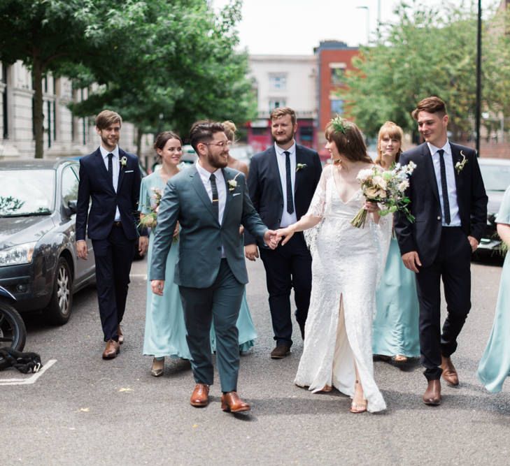 Bride in Grace Loves Lace Verdelle Dress with V-Neck, Low Square Back, Mid-Thigh Front Split and Lace Cape | Foliage Hair Crown | Gold Flat Gianvito Rossi Sandals | Groom in Blue Reiss Suit with Brown Grenson Shoes | Bridesmaids in Mismatched Marine Green Rewritten Dresses | Pastel Wedding Bouquets | Groomsmen in Navy Suits with Le Colonel Moutarde Ties | Neon Sign and Marine Green Rewritten Bridesmaids Dresses with Lace Cape Grace Loves Lace Gown | Alain Mbouche Photography