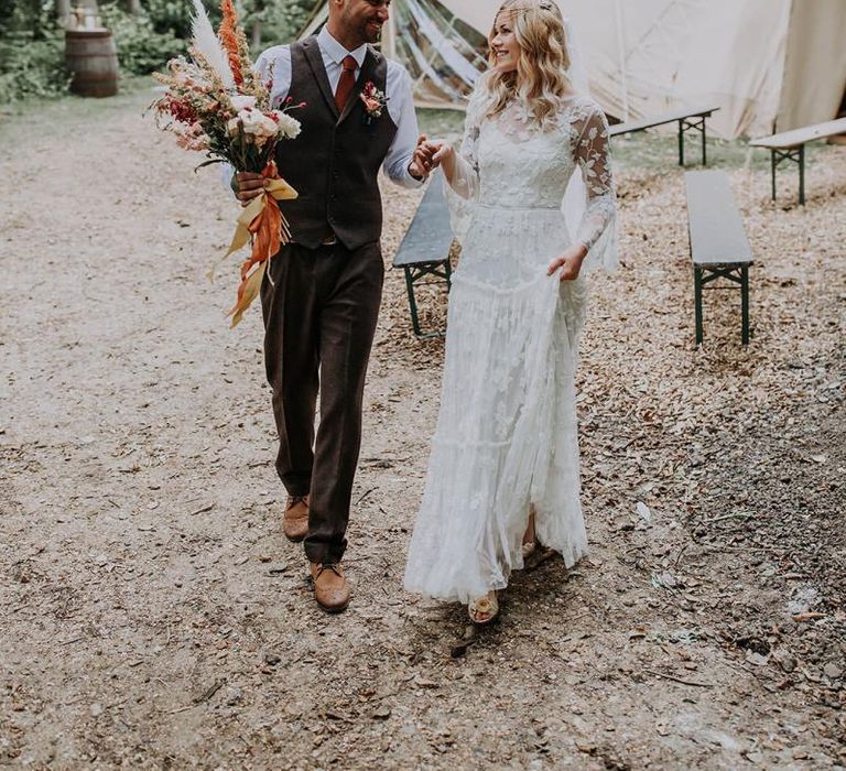 Boho Bride in Lace Wedding Dress with Long Sleeves and Groom in Wool Waistcoat Holding a Bouquet