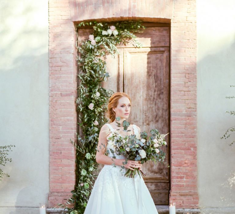 Bride Standing by a Floral Doorway in Lace Sorrisi di Gioia Wedding Dress Holding in a Eucalyptus  &amp; Pink Rose Bouquet