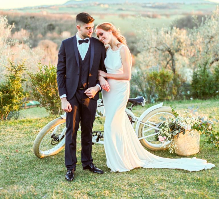 Bride in Lace Wedding Dress and Groom in Tuxedo Embracing by a Vintage Bicycle