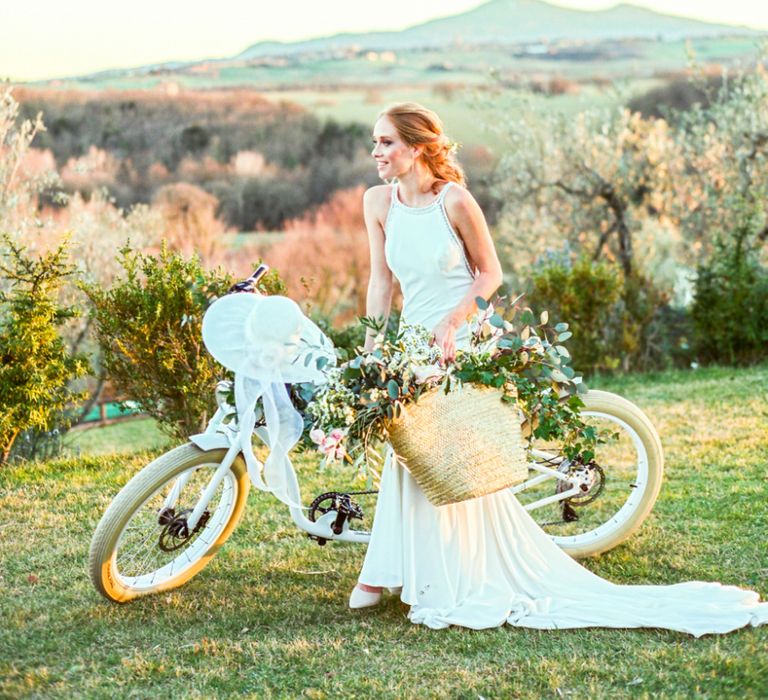 Bride in Lace Wedding Dress Standing Next to  a Vintage Bicycle Holding a Straw Bag of Flowers