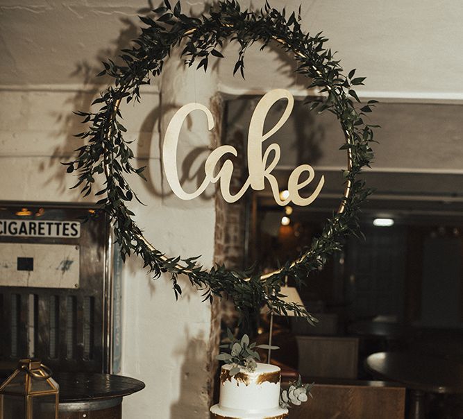 Two Tier Wedding Cake with Foil Decor and Hoop Cake Sign Covered in Foliage