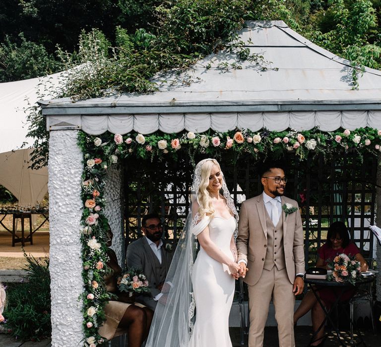 Bride and Groom Standing Under The Flower Covered Pergola at Their Outdoor Wedding Ceremony