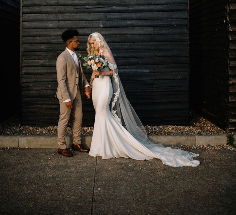 Bride in Pronovias Wedding Dress and Groom in Beige Moss Bros. Suit  Standing in Front of a  Shed Backdrop