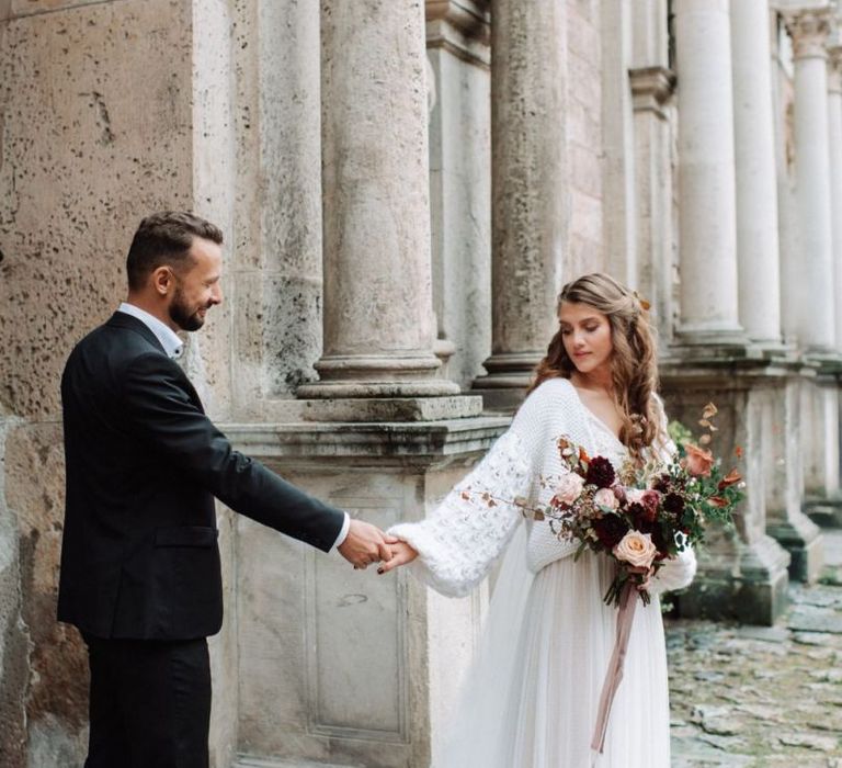 Bride in Katya Katya Wedding Dress with Chiffon Skirt and Knitted Coverup and Groom in Black Suit Holding Hands