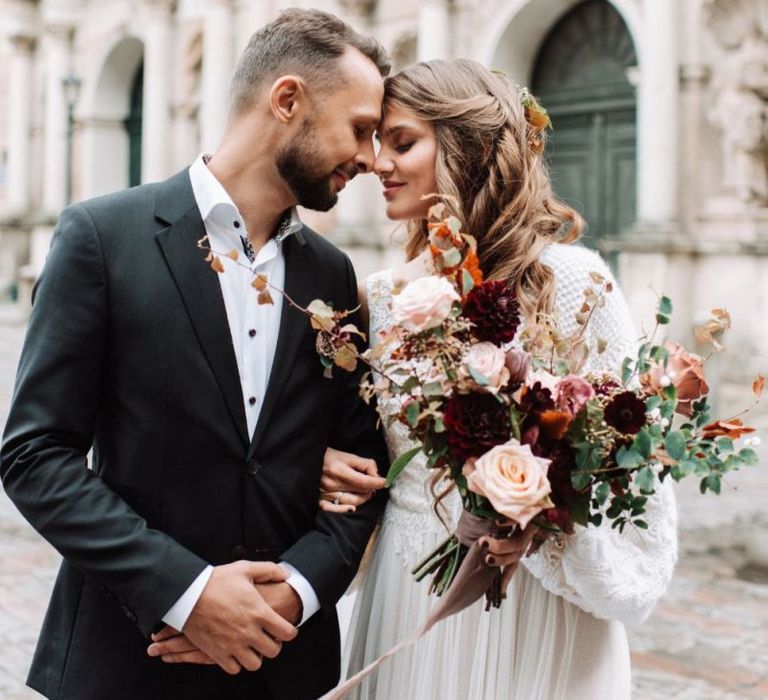 Groom in Black Suit and White Skirt and Bride in Knitted Coverup Holding a Deep Red and Pink Wedding Bouquet