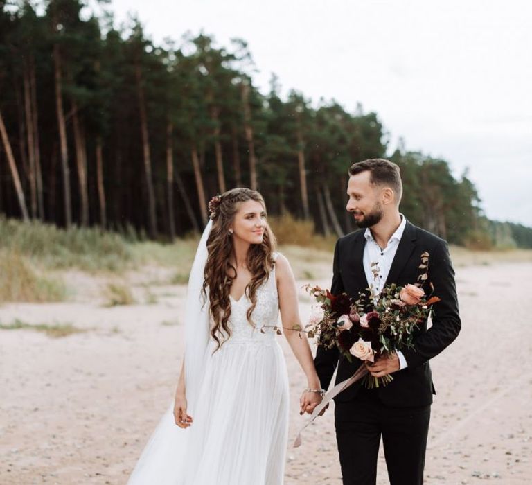 Bride in Fay Katya Katya Wedding Dress with Long Wavy Hair and Groom in Black Suit Holding Hands Walking Along The Beach