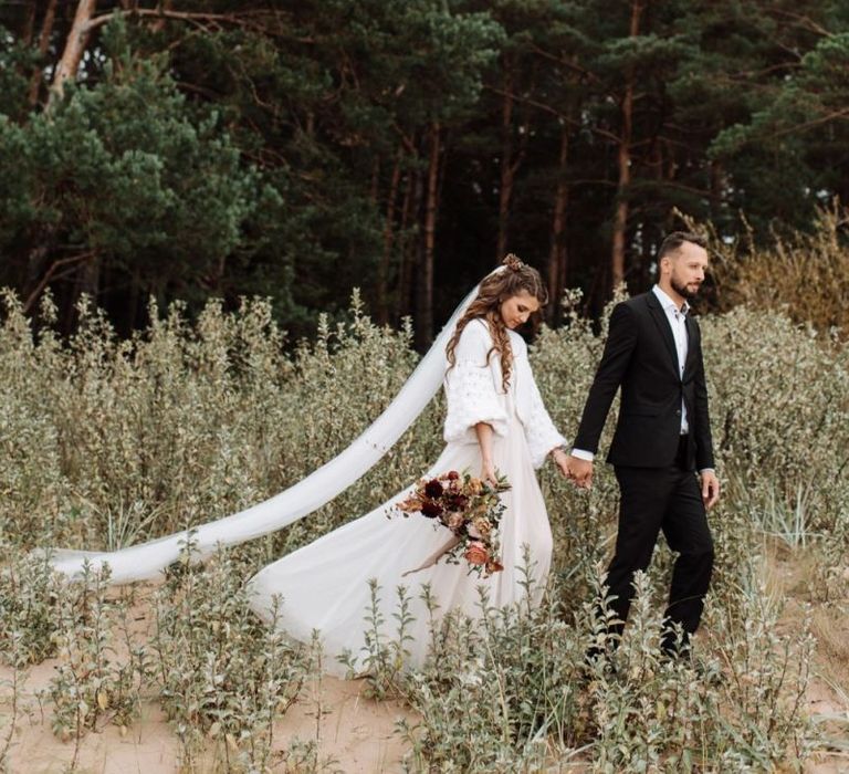 Groom in Black Suit Holding His Brides Hand in a Katya Katya Wedding Dress with Wool Coverup and Cathedral Length Veil