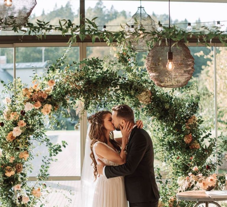 Bride in Katya Katya Wedding Dress and Groom in Black Suit Kissing at the Floral Moon Gate Altar