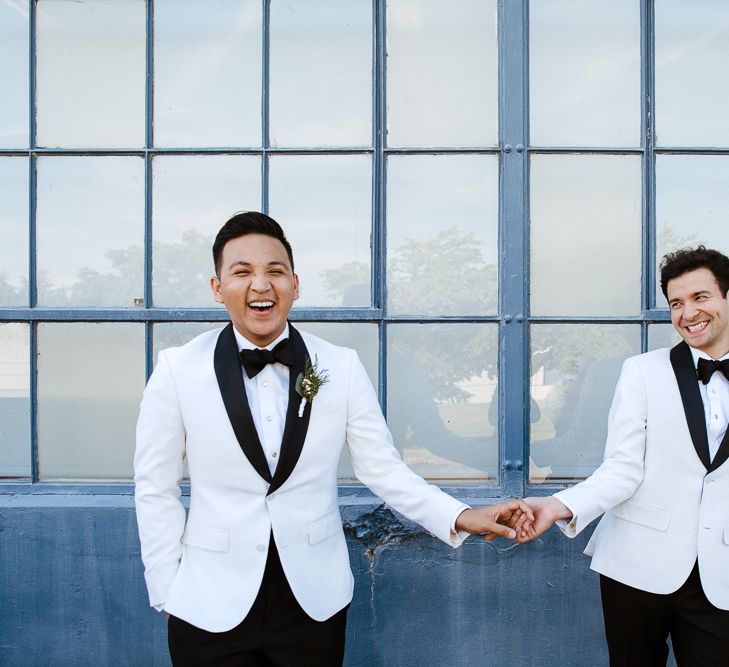 Grooms in white dinner jackets holding hands outside their industrial wedding venue