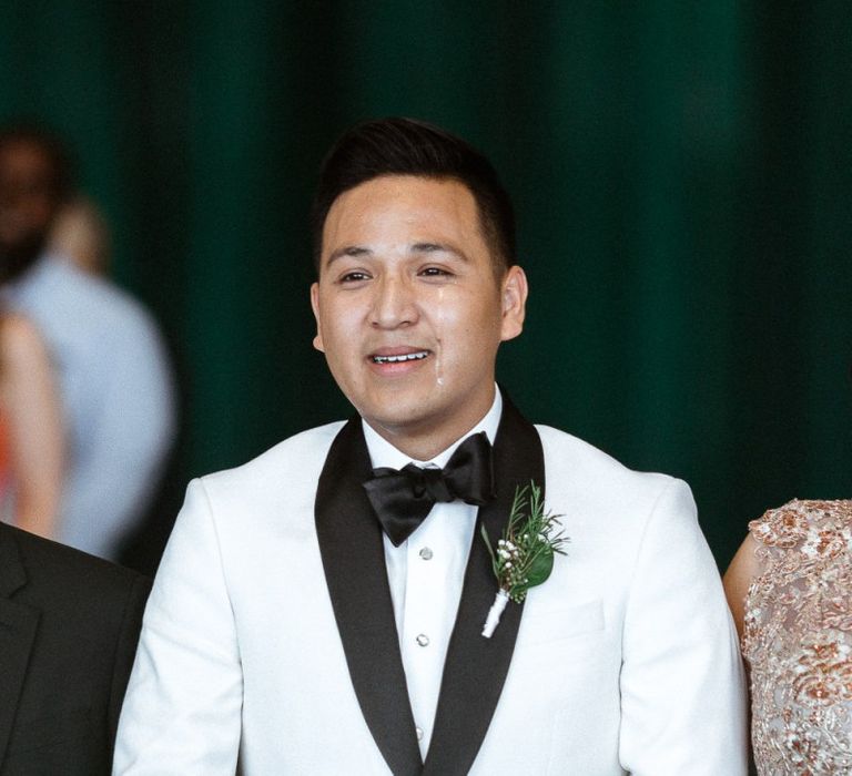 Emotional groom in a white tuxedo jacket walking down the aisle with his parents