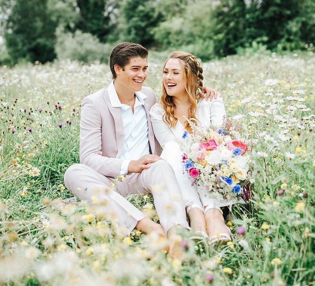 Groom in pink suit and boho bride sitting in a flower filled meadow