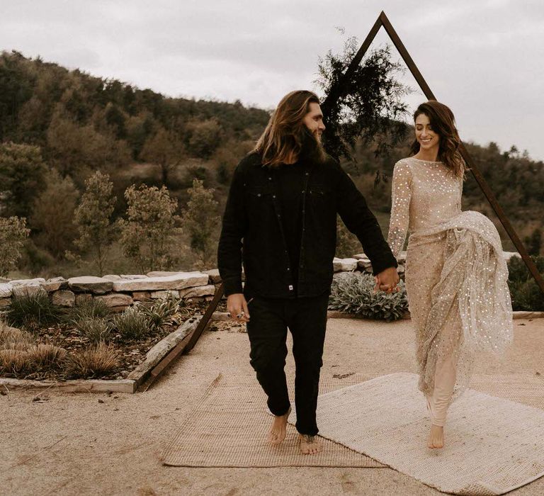 Bride in Gold Shimmering Wedding Dress and Groom in Black Outfit Standing in Front of Triangle Construction.