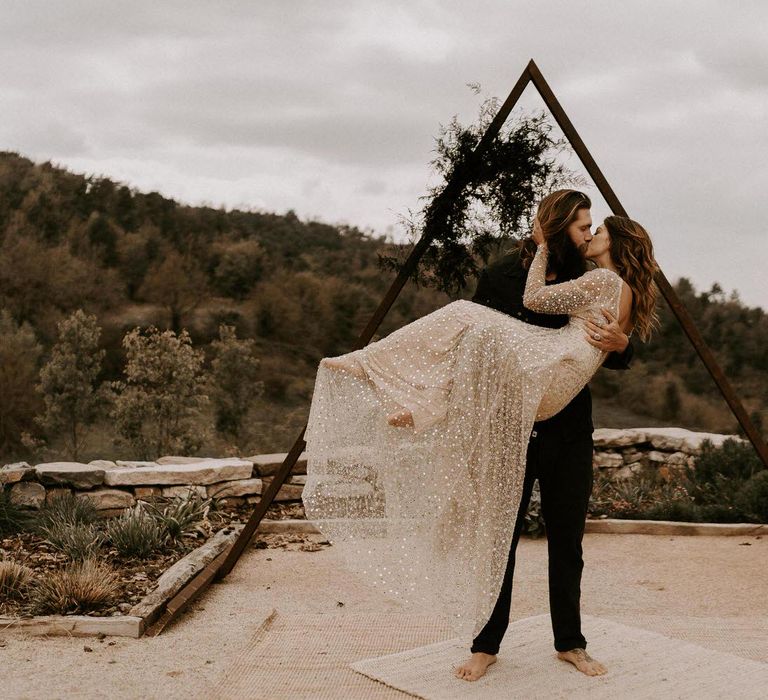 Bearded Groom in Black Outfit Picking Up his Bride in Shimmering Gold Wedding Dress in Front of Triangle Frame