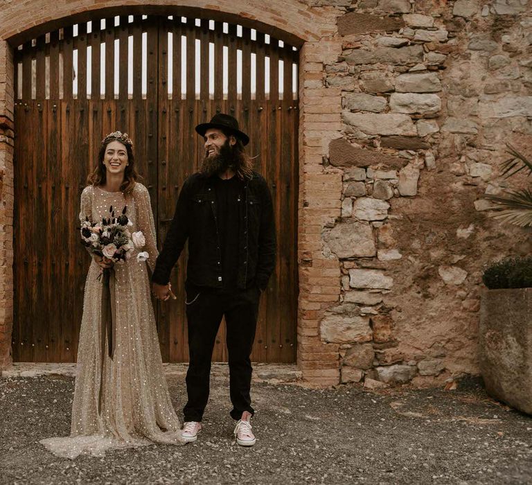 Bride in Shimmering Gold Wedding Dress Holding Wedding Bouquet Tied with Ribbons and Groom in Black Outfit and Fedora Hat