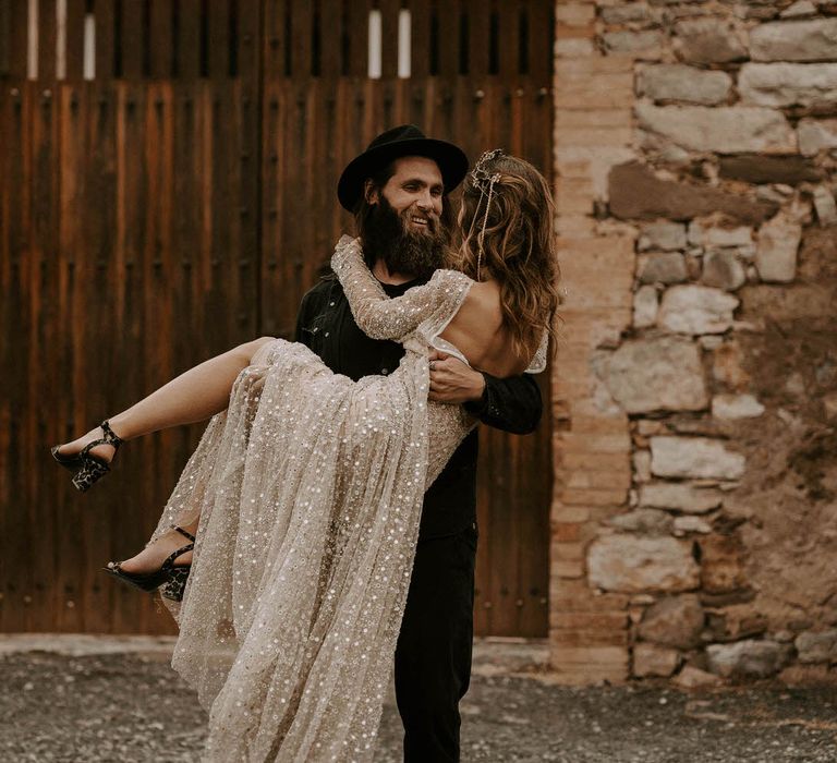 Groom in Black Outfit and Fedora Hat Picking Up His Bride in Shimmering Gold Wedding Dress