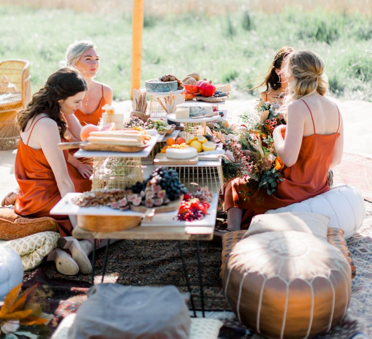 Bridesmaids in Rust Coloured Dresses Enjoying a Grazing Table on Moroccan Pouffe Under a Naked Tipi