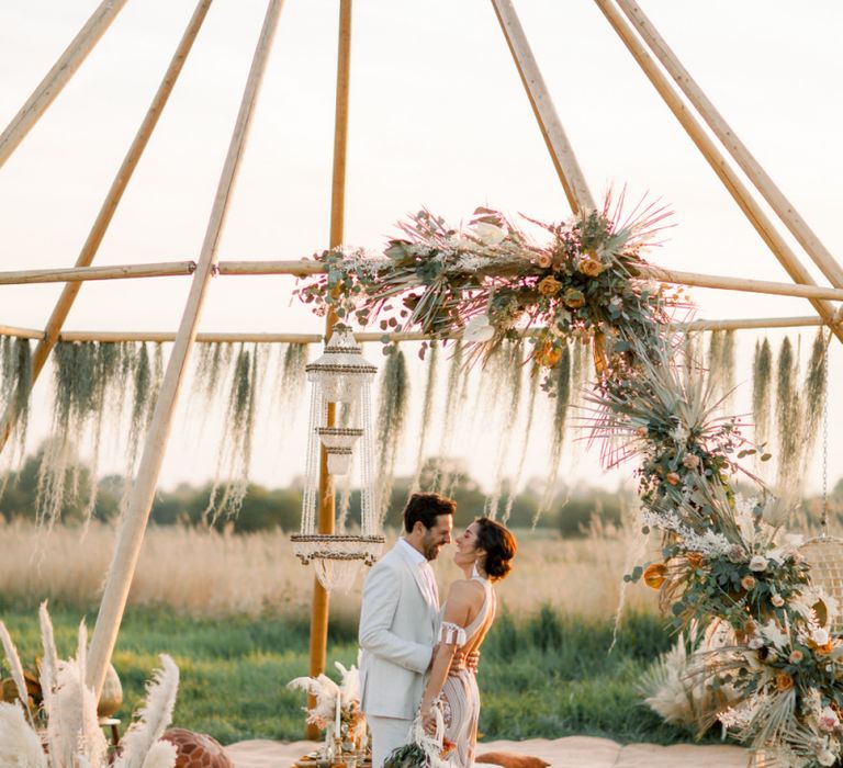 Bride in Rue De Seine Boho Wedding Dress and Groom in Light Suit Laughing in Front of a Naked Tipi Chillout Area