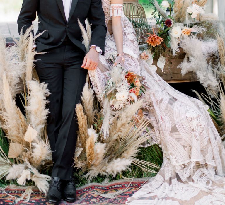 Bride in Rue de Seine Boho Wedding Dress and Groom in Tuxedo Sitting amongst Dried Flower Arrangements