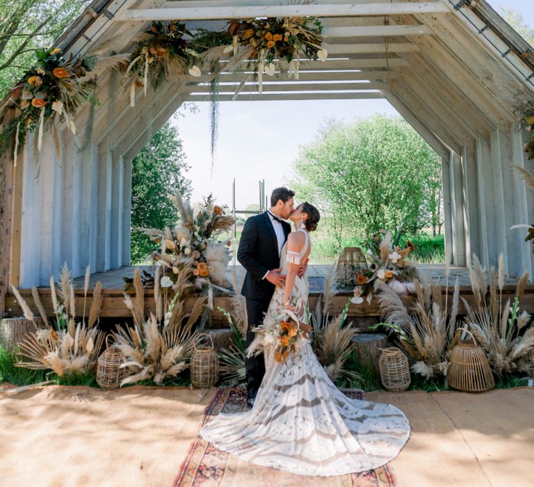 Bride in Rue De Seine Boho Wedding Dress and Groom in Tuxedo Kissing at Their Wooden Hut Outdoor Wedding Ceremony