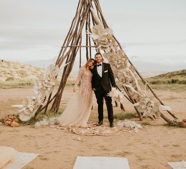 Boho bride and groom standing in from of a naked tipi structure