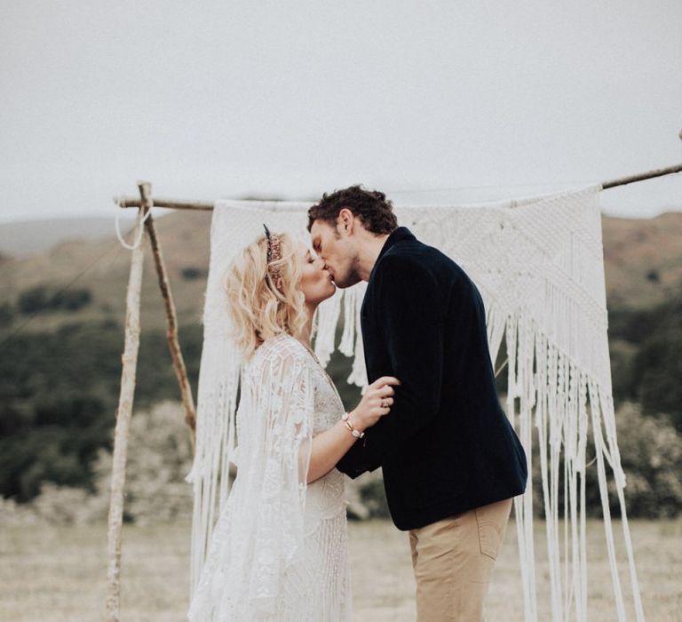 Boho Bride in Lace Rue de Seine Wedding Dress and Crown and Groom in Blazer and Chinos Kissing at the Macrame Altar