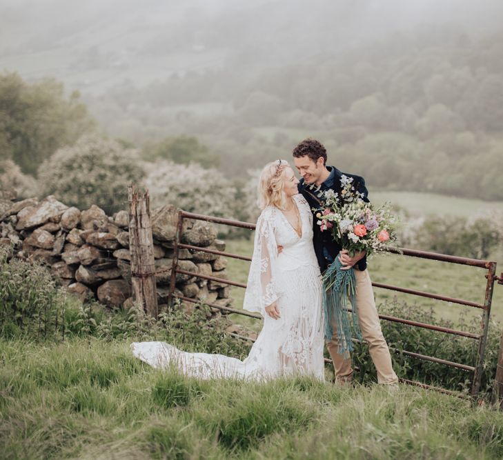 Boho Bride in Rue de Seine Wedding Dress and Groom in Chinos and Blazer Embracing in a Field