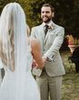 Groom wears classic stone beige linen suit, white shirt and wool green tie alongside a floral buttonhole
