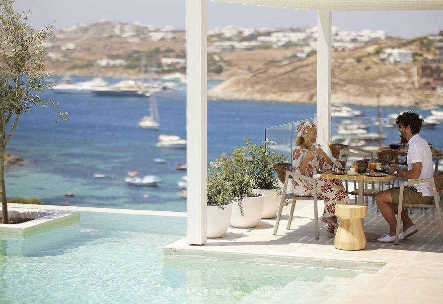 A couple enjoying breakfast at Once in Mykonos, seated on a shaded terrace with panoramic views of the Aegean Sea and anchored boats